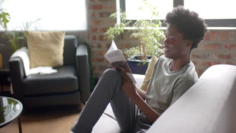 hombre afroamericano con parches debajo de los ojos leyendo un libro en el sofá en casa, en cámara lenta