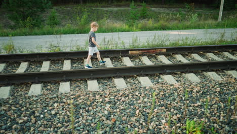 side view of young boy in gray t-shirt and light shorts walking confidently on rail track through quiet countryside with gravel bed, overgrown grass, and dirt road