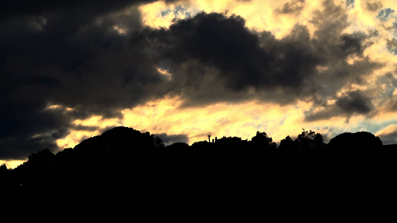 Dark clouds drift over a glowing yellow sky above a hilltop