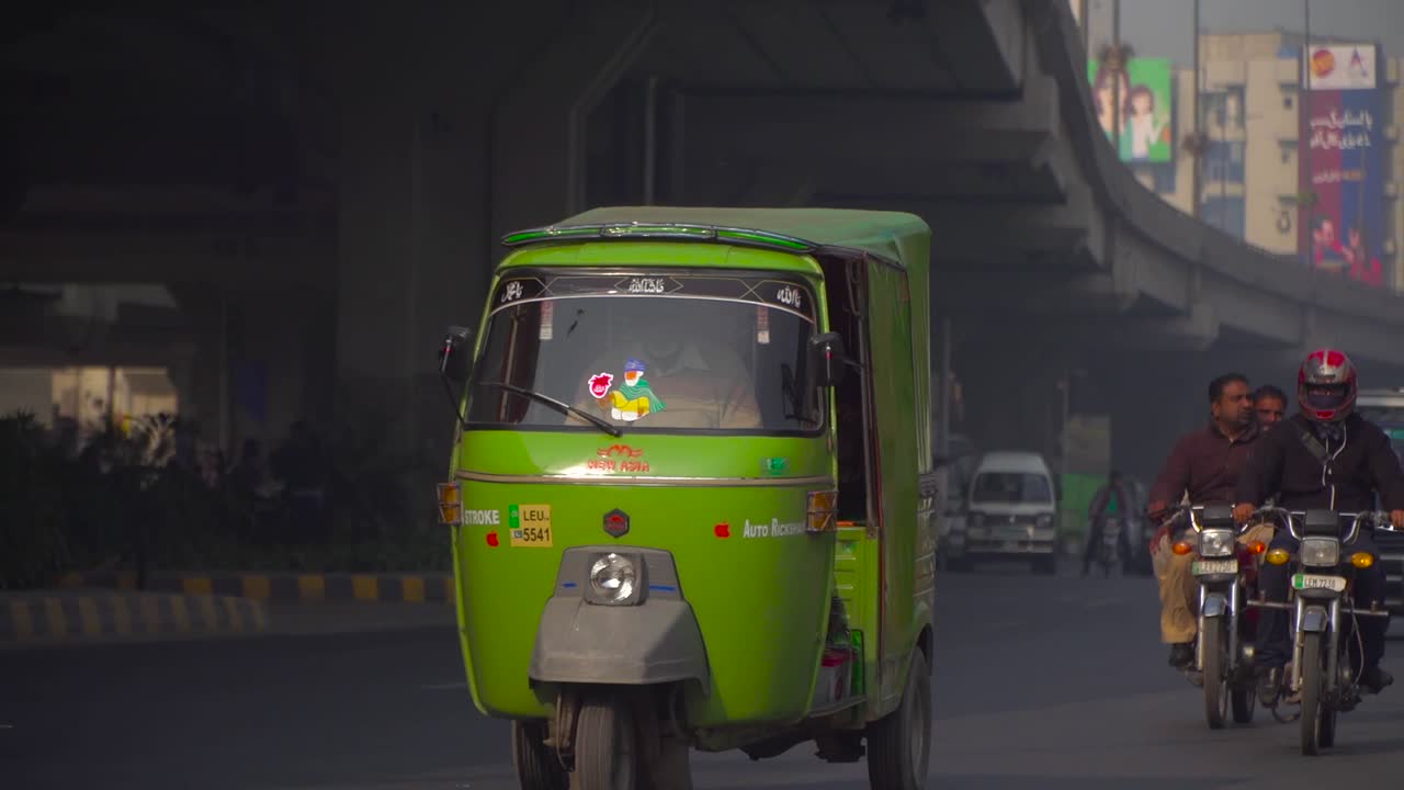 Traffic close up at the road under the fly over, passing motor bikes and cars, Buses, rickshaw`s, Billboards and buildings in the background