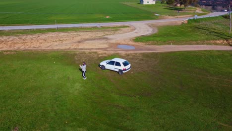 aerial view man piloting a drone near to a small car surrounded by green meadows and countryside