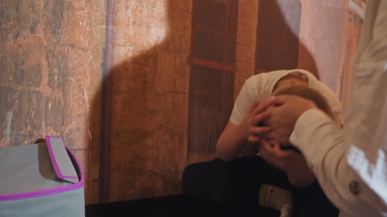 Madre e figlio bianchi giocano in casa, con la madre dolce che consola e solletica il bambino vicino a una parete texturizzata con un'ombra di foglie. La calda luce solare proietta una silhouette, mentre un gatto soriano osserva nelle vicinanze e uno zaino è a terra.