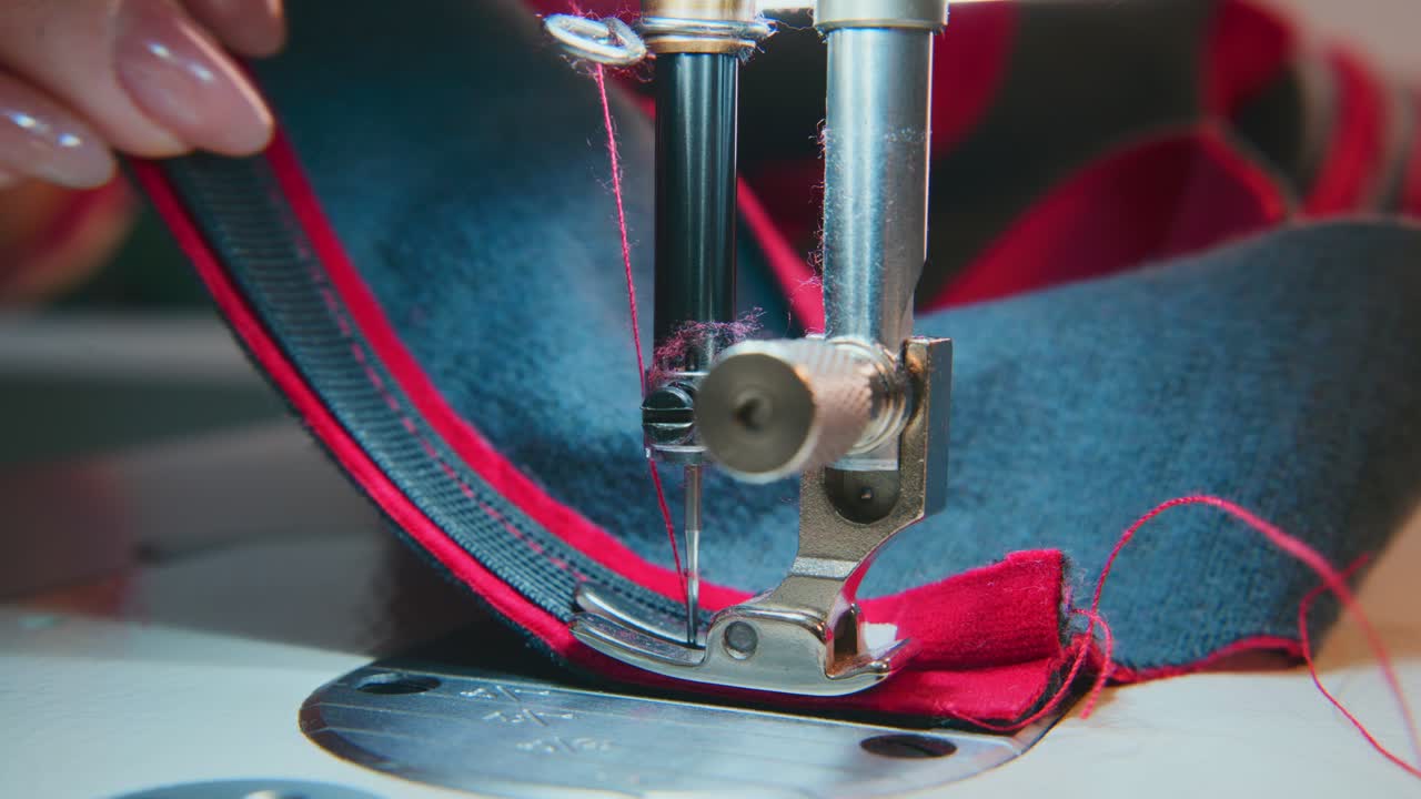 Nærbillede af hænder, der fører stof under symaskinens nål. Fingre justerer forsigtigt materialet for præcise syninger. Rød tråd kontrasterer med mørkt stof.