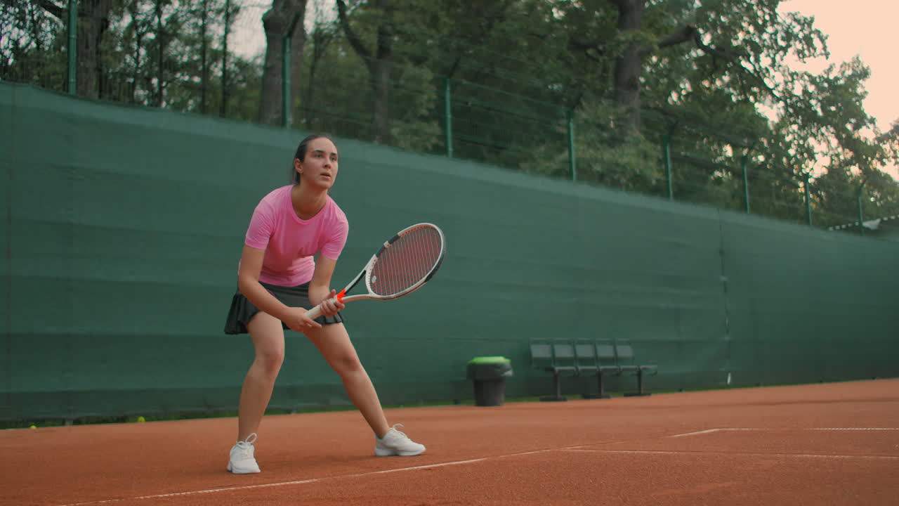 한 젊은 여성이 해가 지는 동안 단단한 표면의 테니스 코트에서 훈련을 받고 포 ⁇  풀  ⁇ 을 기다리고 있습니다.