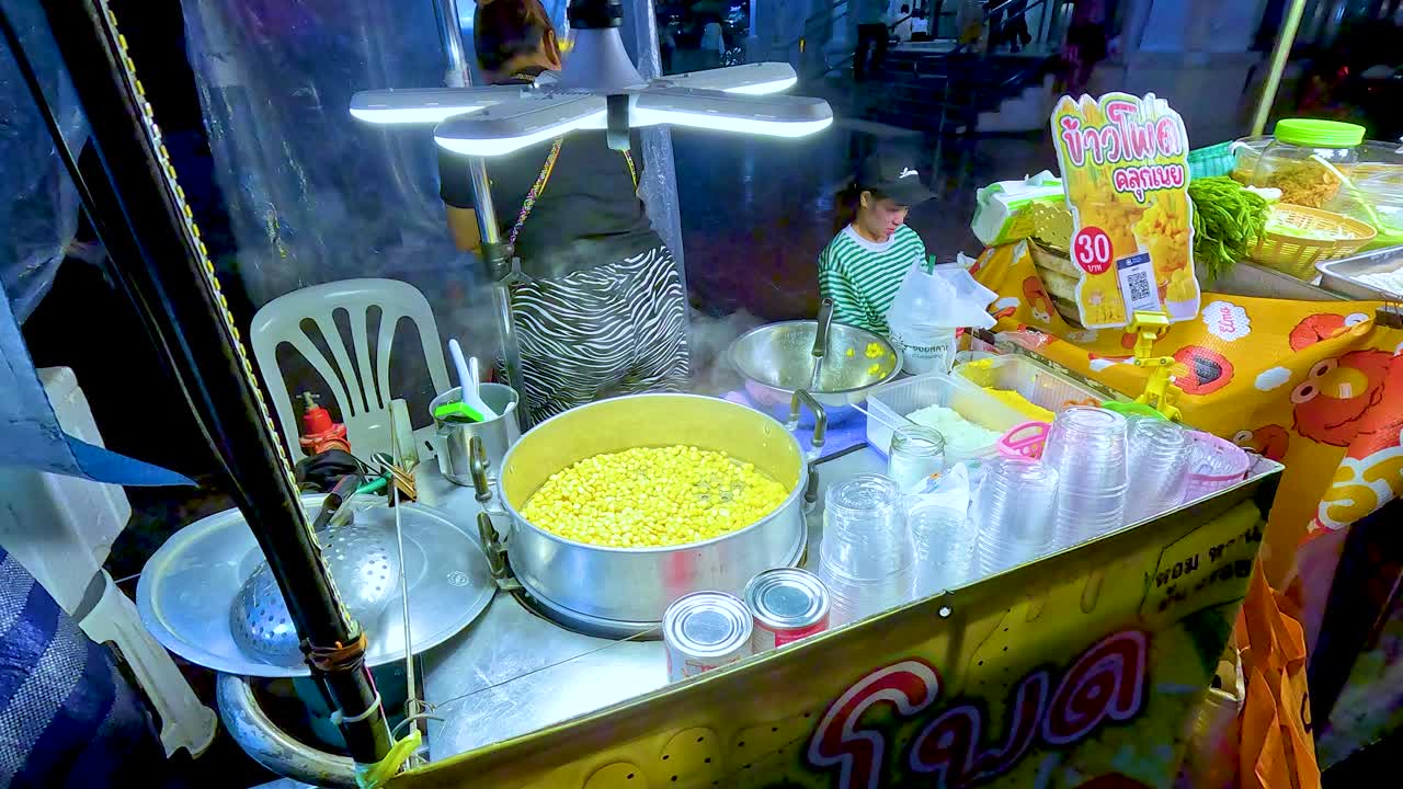 Female vendor prepares steamed corn at colorful night market stall, overhead view, artificial lighting