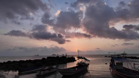 Stunning-sunset-in-Koh-Samui-Thailand,-displaying-silhouettes-of-clouds,-rocks,-long-tail-boats,-and-fishing-boat-on-shore-while-the-setting-sun-is-covered-by-clouds