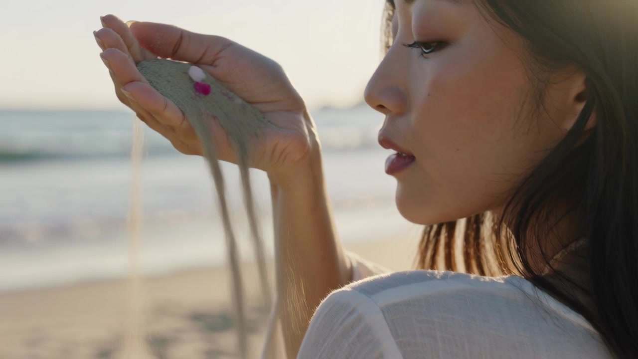 Mano che si muove, donna che osserva la sabbia scorrere sulla spiaggia, con top trasparente e unghie rosa, spazio per il testo.
