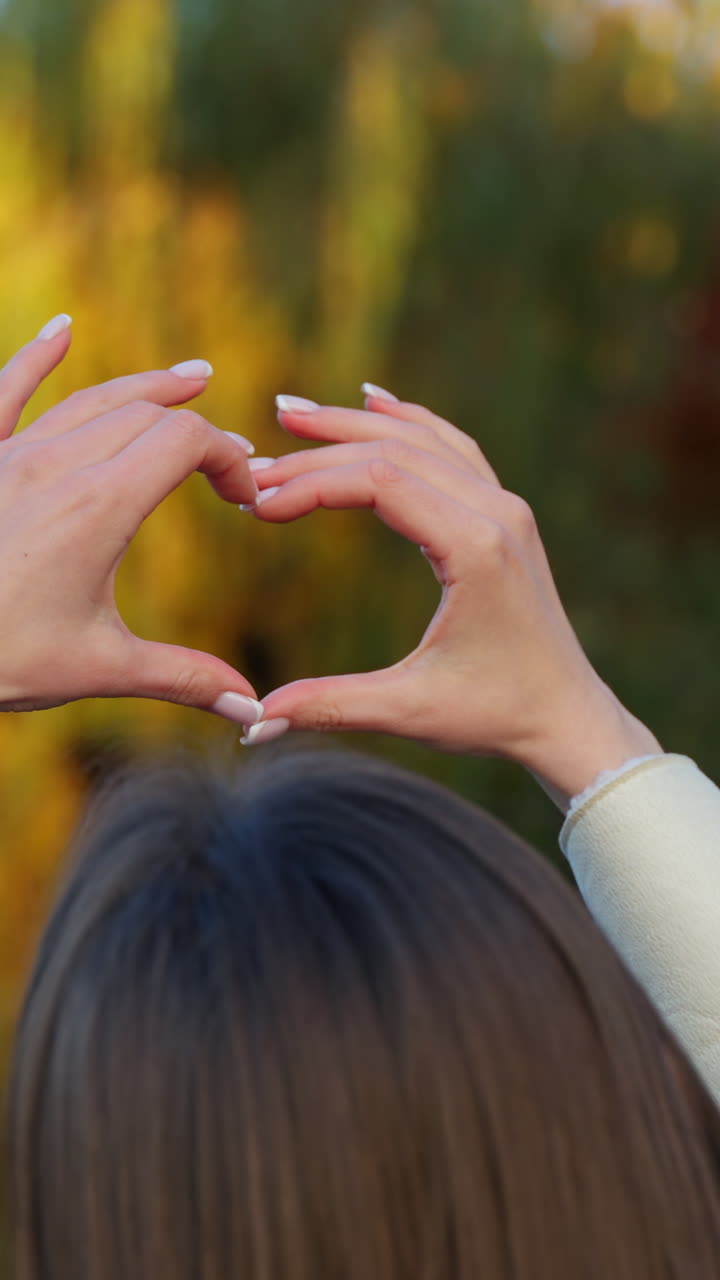 Unknown brunette outdoors holds up her hands to make the heart sign. Beautiful sunny autumn colorful trees at backdrop in blur. Vertical video