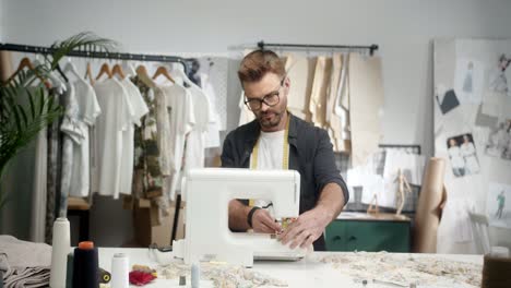 man tailor in glasses sitting at the table with a sewing machine in his workshop and smiling cheerfully to the camera