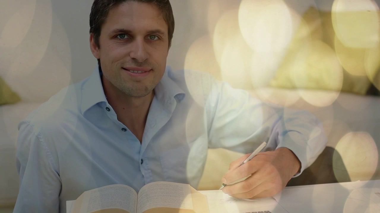 Homem inclinando-se para frente, rabiscando com caneta, virando páginas estudando livro aberto com sobreposição de bokeh quente.