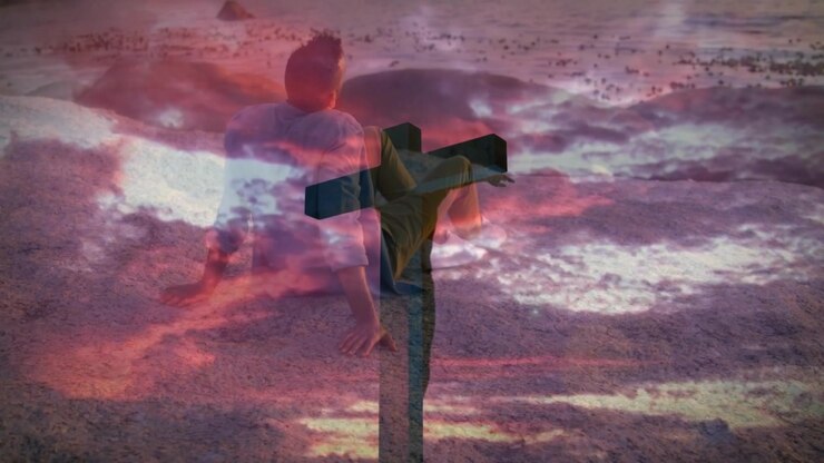 Male seating sandy shore during meditation causing crucifix fading and sky shifting into pink hues