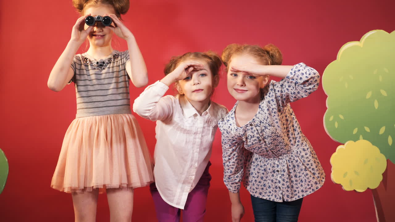 three young girlfriend are considering everything around themselves on the red background. One of little girl in dress is looking through binoculars