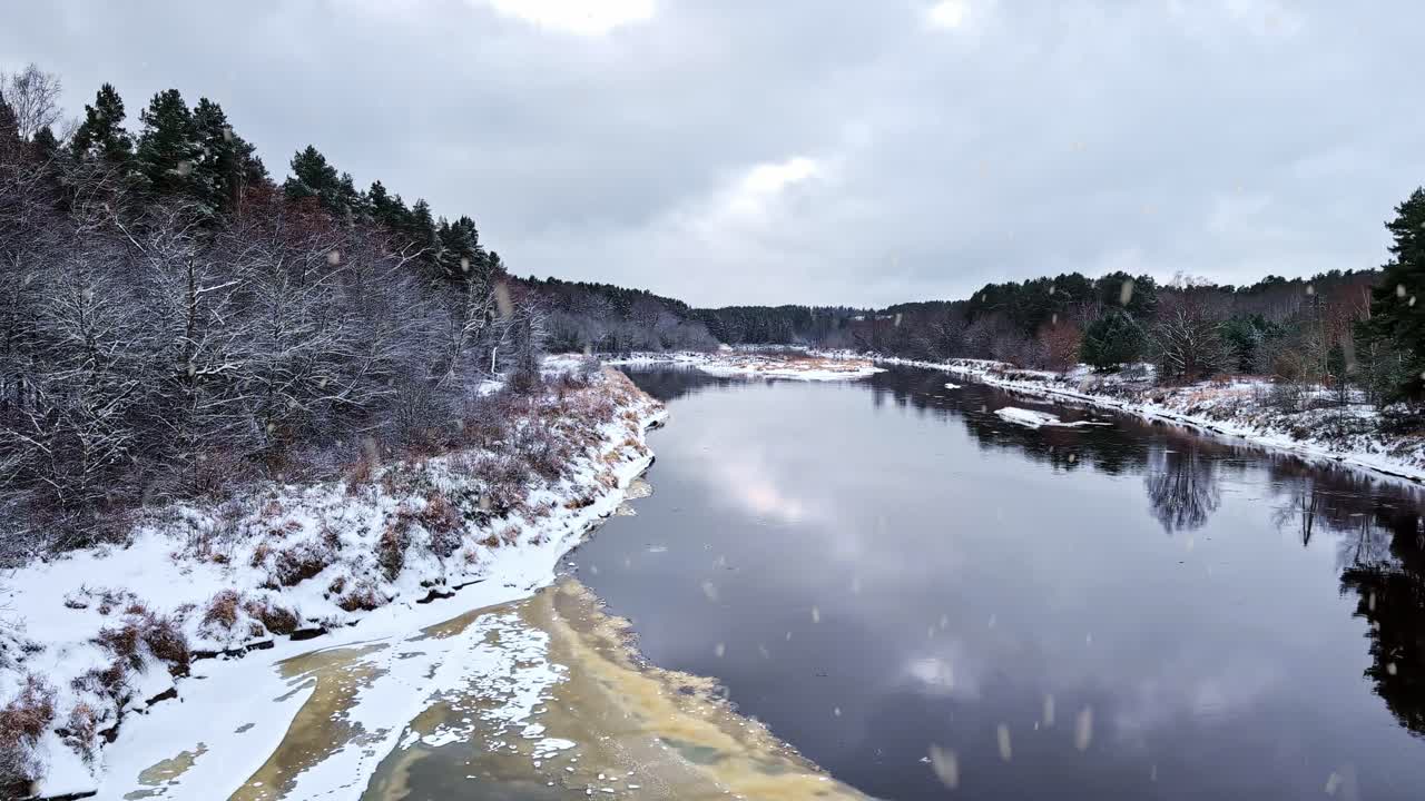 Osittain jäätynyt Gauja-joki mutkittelee lumisateessa lumipeitteisten rantojen ja puiden lomitse pilvisen taivaan alla.