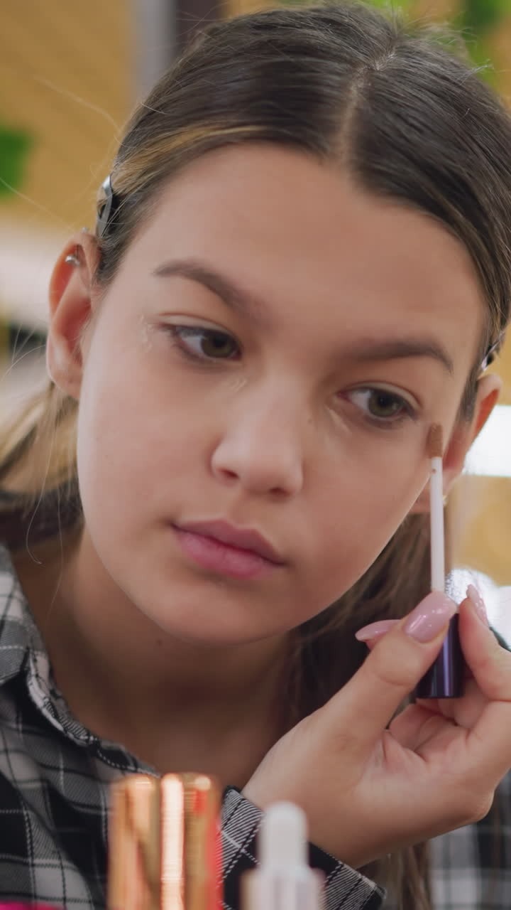 Primer plano de una adolescente aplicándose corrector líquido debajo de los ojos, con una mujer usando cuidadosamente un aplicador de varita frente al espejo, iluminación cálida de estudio, expresión tranquila y concentrada, uñas impecables, productos de belleza