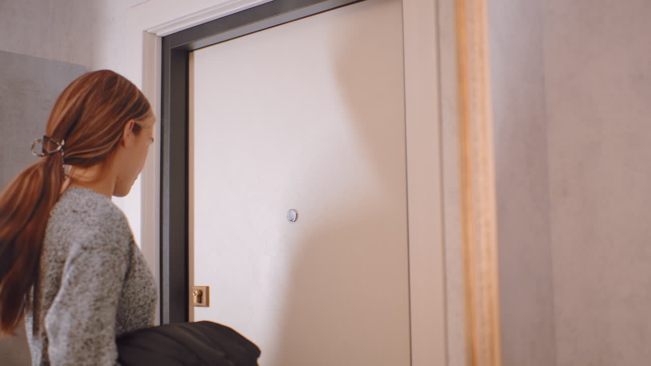 Woman in gray sweater holding black jacket leave house, standing near closed beige door with focused expression, long hair tied with clip, natural indoor light