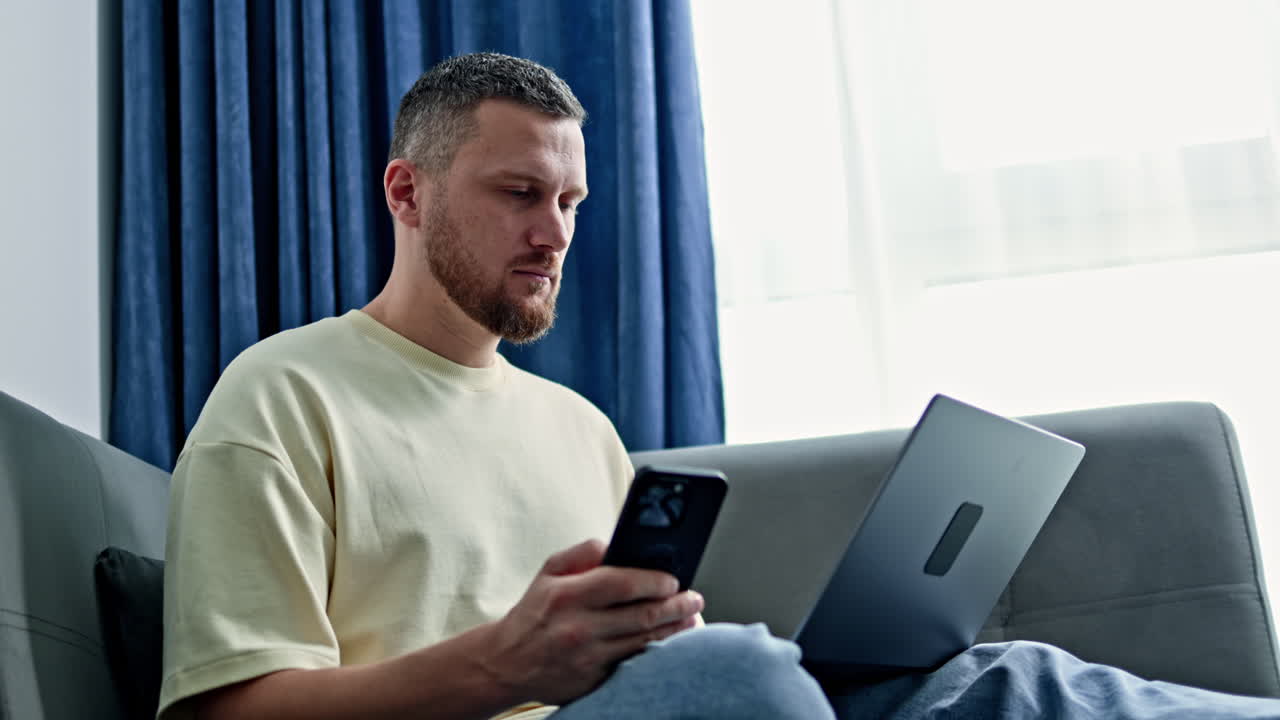 Man sitting on couch using smartphone while working on laptop in a bright room with curtains in the background during daytime