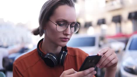 blonde pretty girl with make up standing on the street and typing on her black cell phone. wearing orange shirt and modern