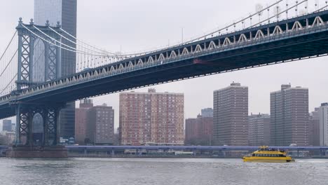 Yellow-water-taxi-passenger-ferry-on-the-Hudson-River-with-cityscape,-traffic-in-background-and-a-bridge-overhead-in-NYC,-New-York-City