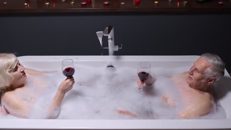 senior couple in love lying in warm bath with bubbles, enjoying relaxation, drinking red wine