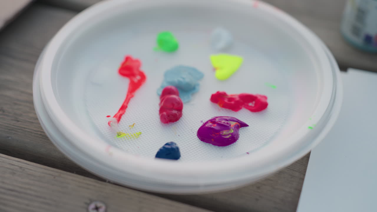 hand holds brush mixing pastel blue acrylic with neon red green yellow pink purple paints on white plastic palette plate on wooden bench by river at sunset outdoor painting session