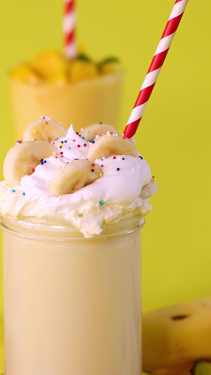 Una mano inserisce una cannuccia a strisce in un cremoso milkshake alla banana guarnito con panna montata e codette colorate.