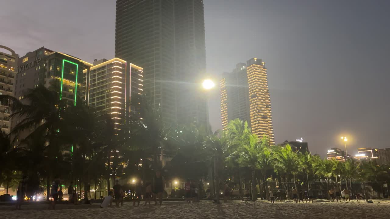Vista noturna de arranha-céus modernos em Da Nang, com edifícios altos iluminados, postes de luz brilhantes e palmeiras contra o céu noturno escuro na área da cidade costeira.