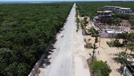 Antena-De-Autos-Conduciendo-Hacia-El-Océano-En-La-Zona-Hotelera-De-Tulum-Mientras-Los-Turistas-Pedalean-En-Bicicleta