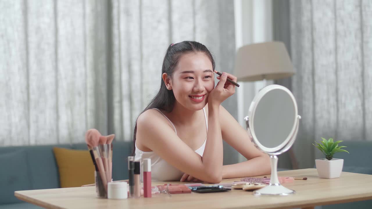 Beautiful Young Asian Woman Looks In The Mirror Brushing Eyebrow And Smiles While Applying Makeup At Home