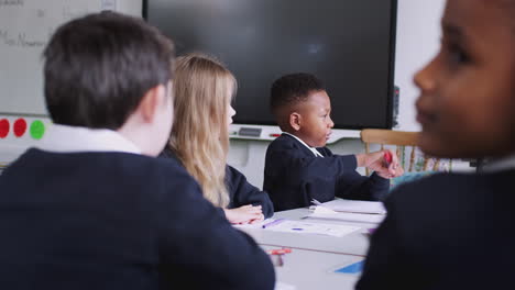 Primary-school-kids-at-desks-in-a-classroom-raising-hands-to-answer-a-question,-back-view,-close-up