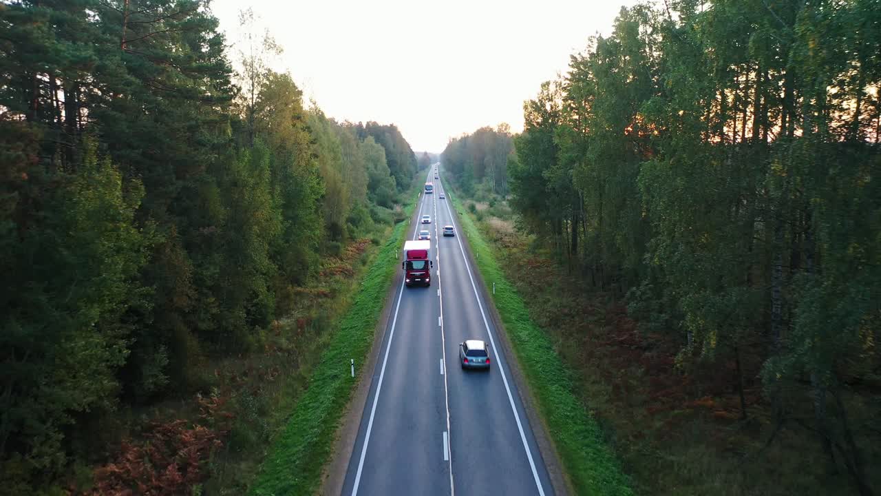 Ujęcie filmowe przedstawia poranną drogę z pojazdami poruszającymi się po wiejskiej Łotwie