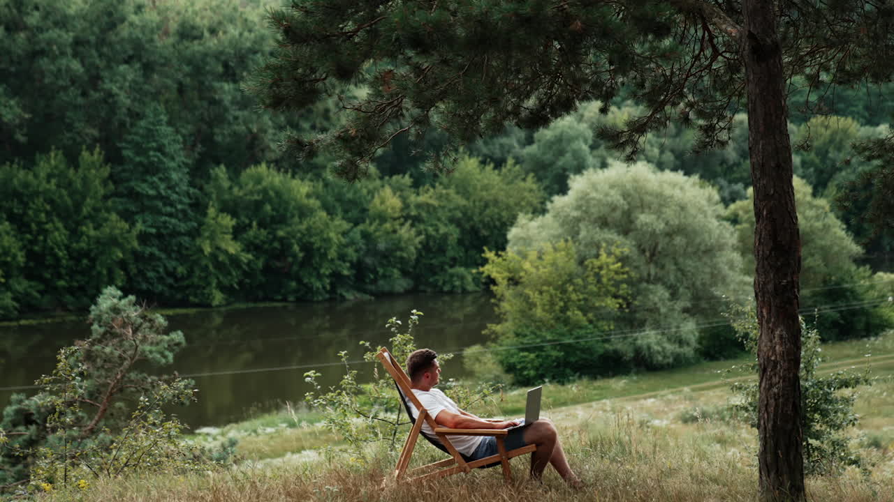 Comfortable workplace in nature remote from office. Freelancer works on laptop sitting in the folding chair. Side view.