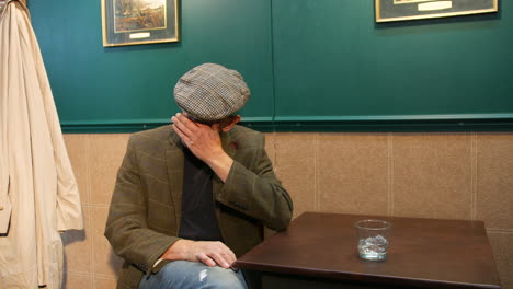 a mature alcoholic man in a flat cap sitting at a table distressed. turning away from a glass of vodka alcohol in a pub in a moment of emotional struggle with addiction