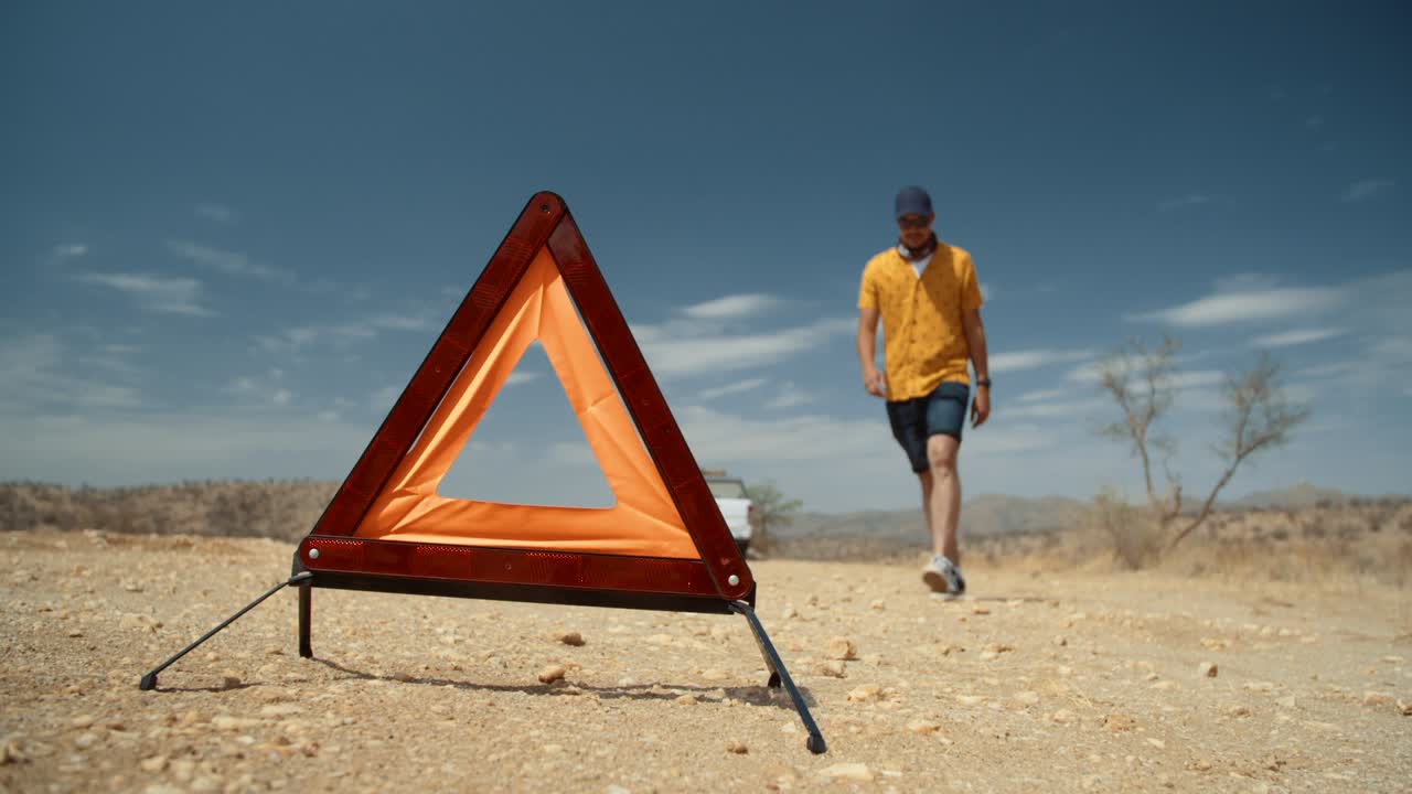 Low angle wide static shot of a Caucasian male tourist in Africa as he removes a reflective emergency road side triangle and proceeds to walk back to his off-road vehicle
