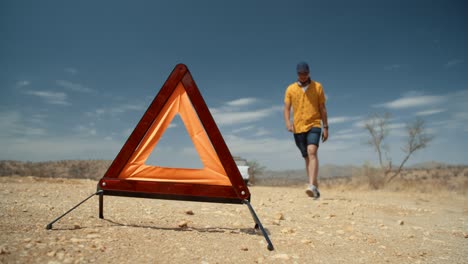 low angle wide static shot of a caucasian male tourist in africa as he removes a reflective emergency road side triangle and proceeds to walk back to his off-road vehicle