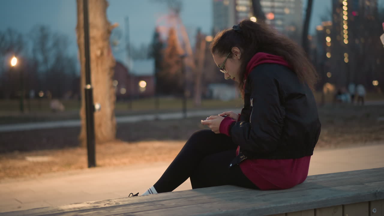 Casual Lady Browsing Social Media Outdoors, Woman In Red Hoodie Enjoying Peaceful Park Evening, Asian Female Seated On Park Bench Reviewing Social Media Content During Quiet Twilight Hours