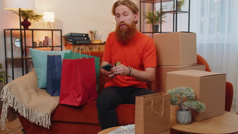 joven adicto a las compras feliz sentado con bolsas de compras haciendo pagos en línea con tarjeta de crédito en casa