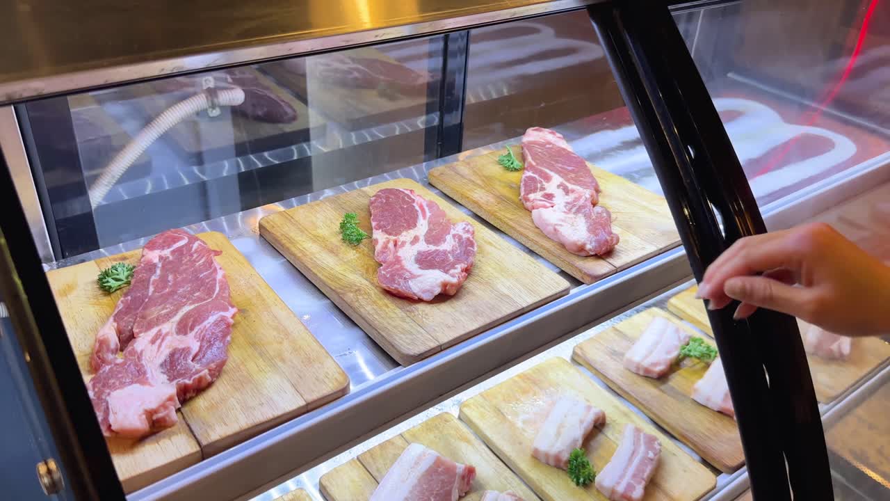 A person selects raw pork cuts from a display case under bright lighting, preparing for a Korean BBQ experience in Bangkok