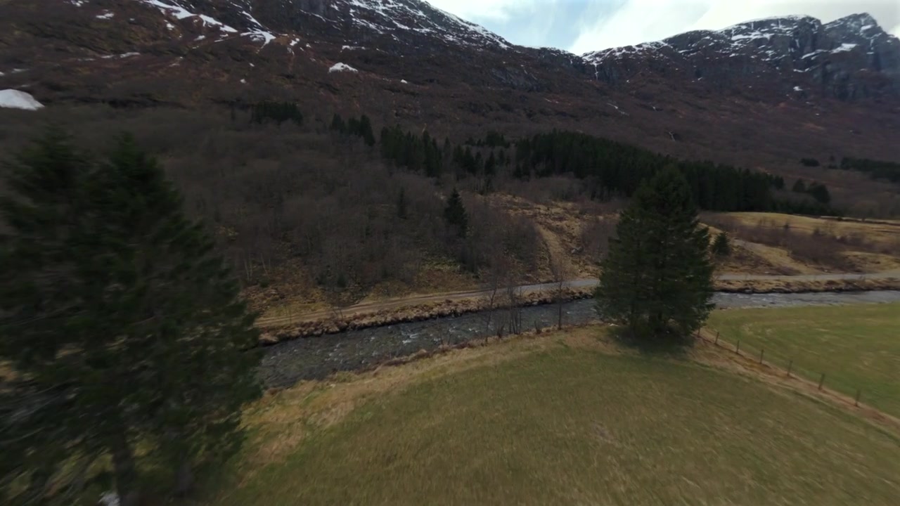 Dron letící vpřed nad travnatým polem směrem k zalesněnému úpatí hory se zasněženými vrcholky v Norsku.