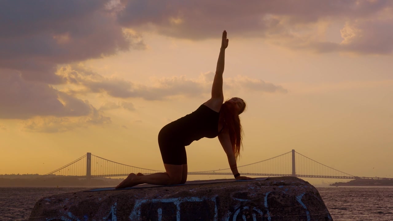 Donna che pratica la posizione yoga del gatto al tramonto a New York. Silhouette femminile che inarca la schiena su una sporgenza di pietra, Ponte di Verrazzano Narrows, cielo dorato, acqua.