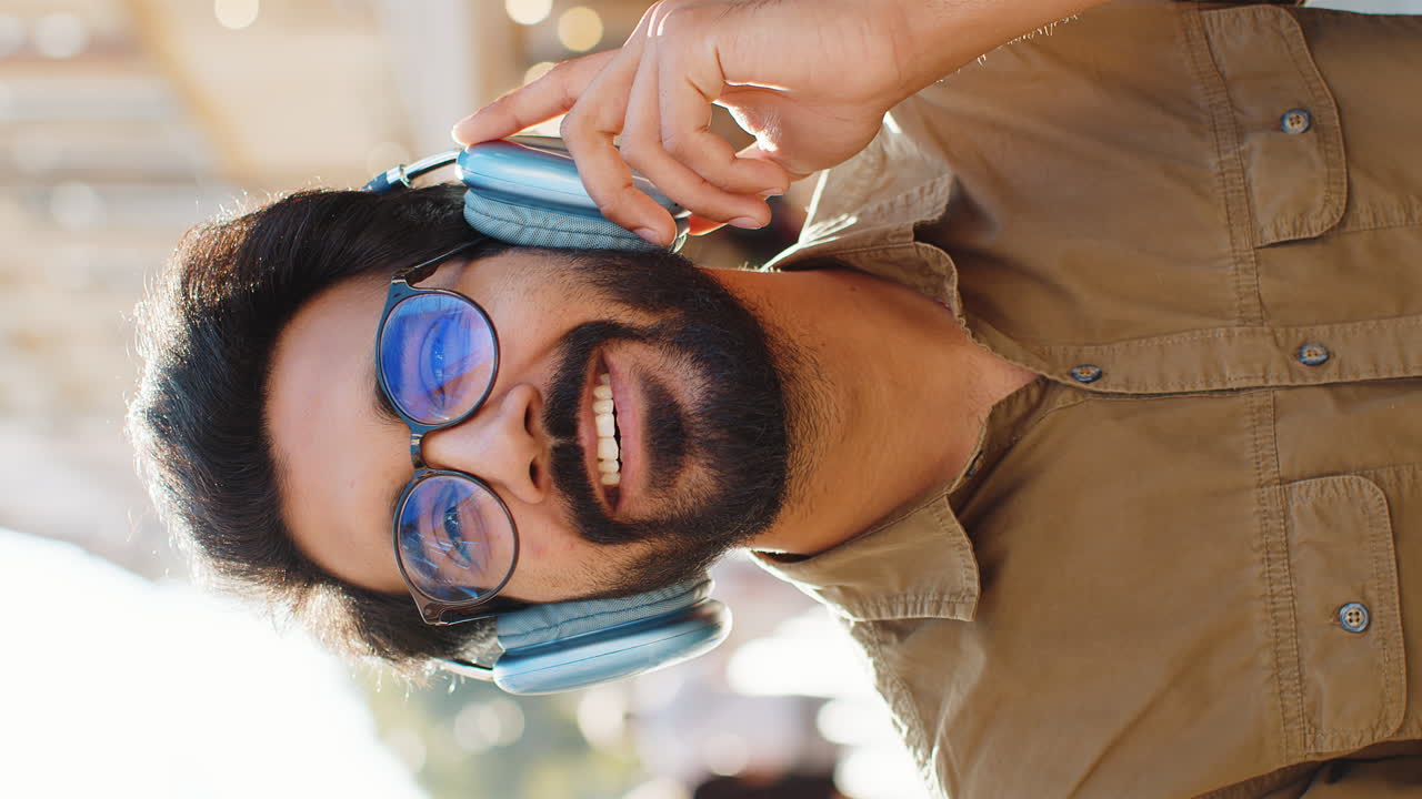 Happy young adult man in wireless headphones choosing listening music dancing outdoors city street