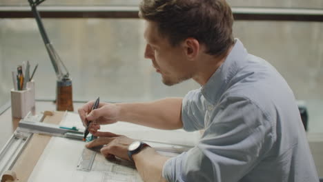 a male architect sitting at a table draws a plan of the building and is engaged in design development sitting in the office in the sitel loft near a large window.