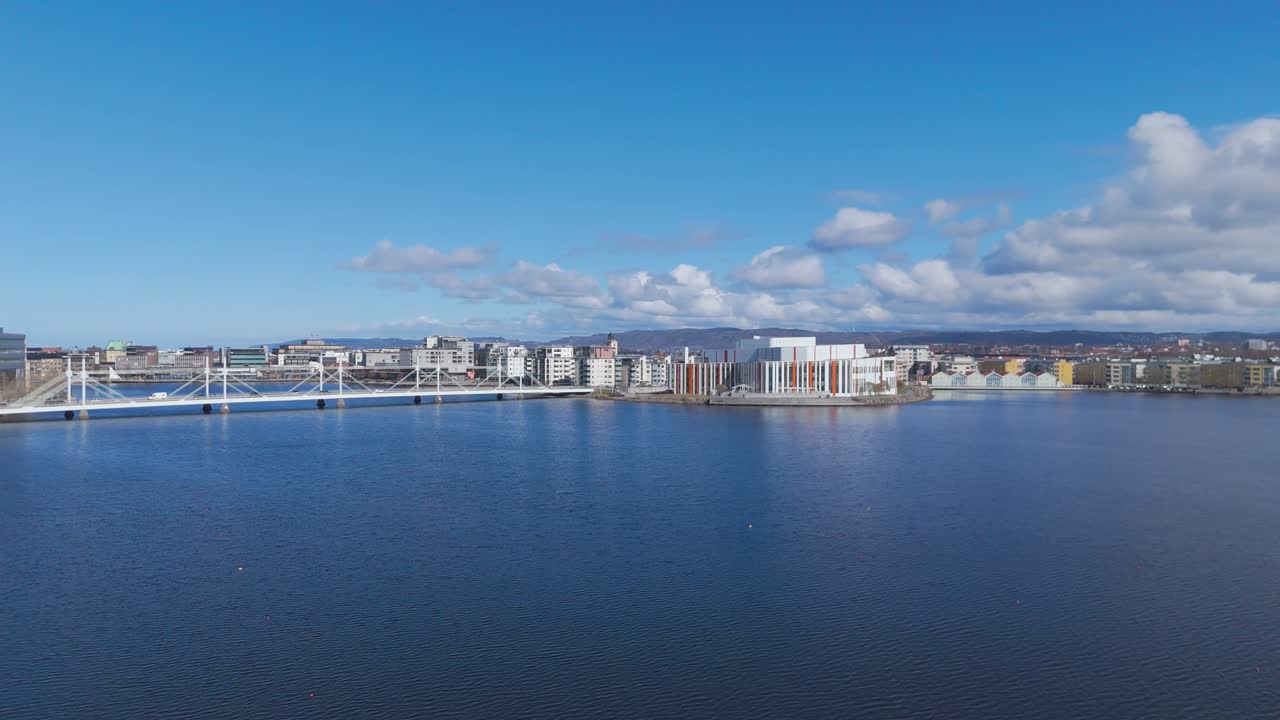 Ampia vista aerea del porto di Bergen, Norvegia, con moderni edifici sul lungomare, mare calmo e colline distanti sotto un cielo azzurro brillante con nuvole sparse, città costiera panoramica