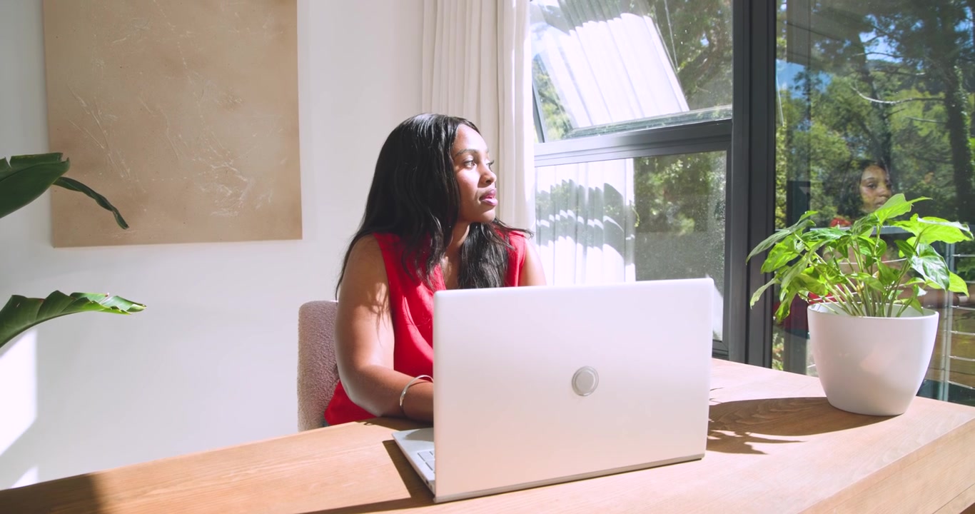 Woman in sleeveless red top typing on laptop at desk, pausing, glancing at outside figure, resuming