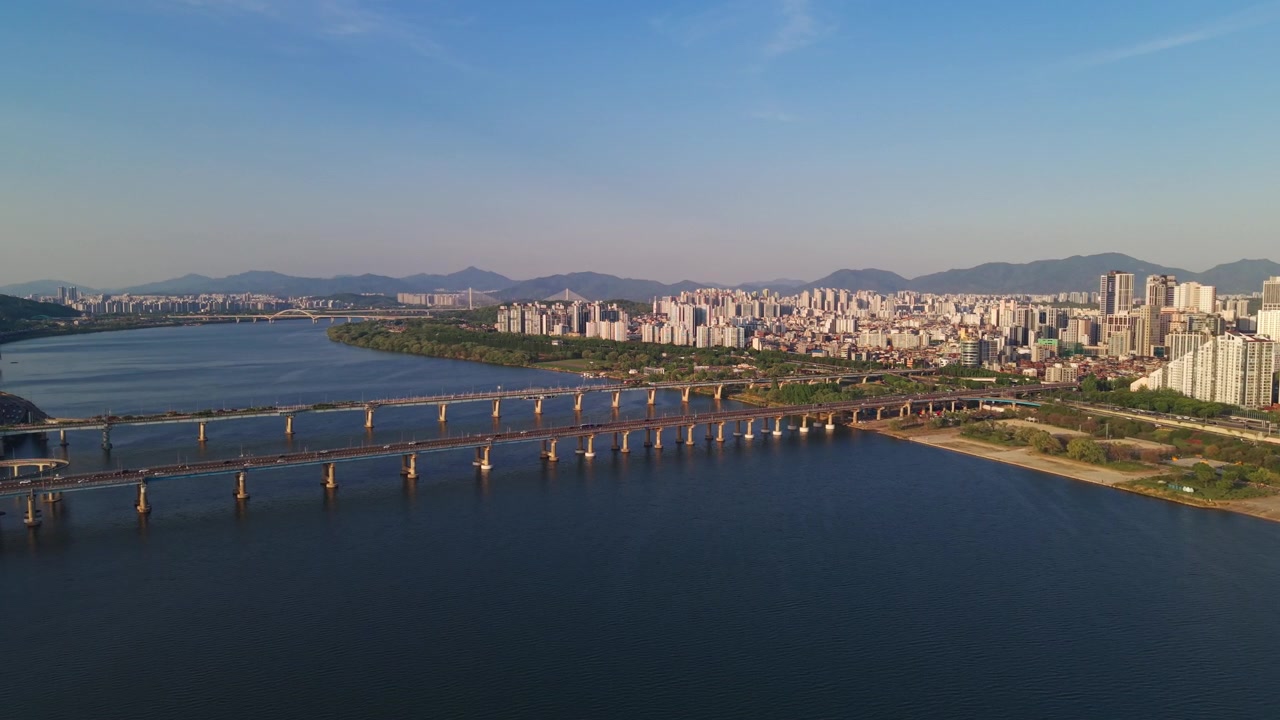 Discesa aerea all'ora d'oro sul fiume Han verso i ponti Gwangjin e Cheonho con il parco Gwangnaru, lo skyline primaverile di Seul e le montagne.