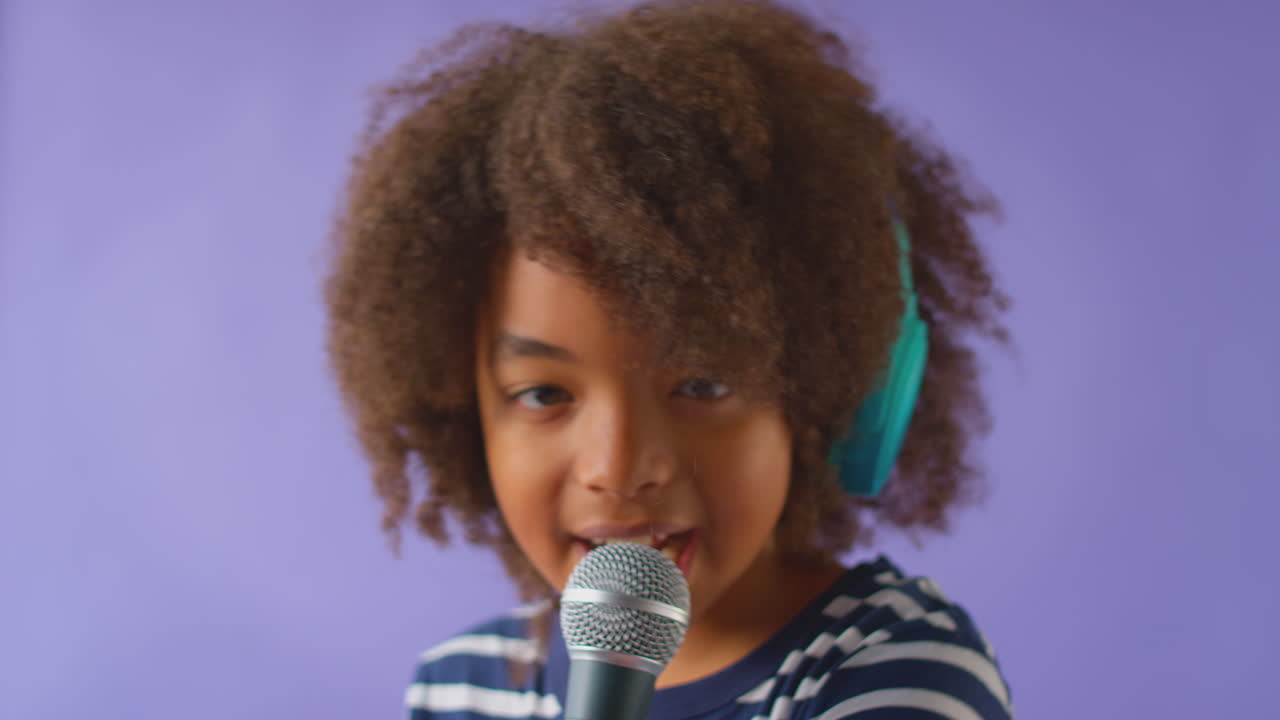 Studio Shot Of Boy Wearing Headphones Singing Karaoke Into Microphone Against Purple Background