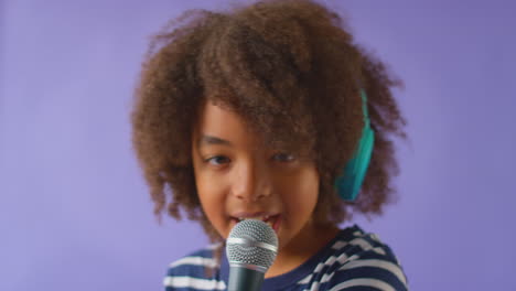 Foto-De-Estudio-De-Un-Niño-Con-Audífonos-Cantando-Karaoke-En-El-Micrófono-Contra-Un-Fondo-Morado