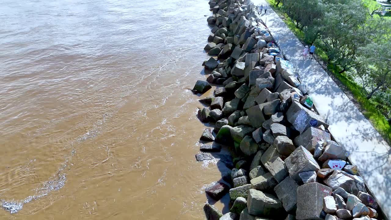 Aerial drone view of geometric rock breakwater, brown river, and tree-lined path in Australia