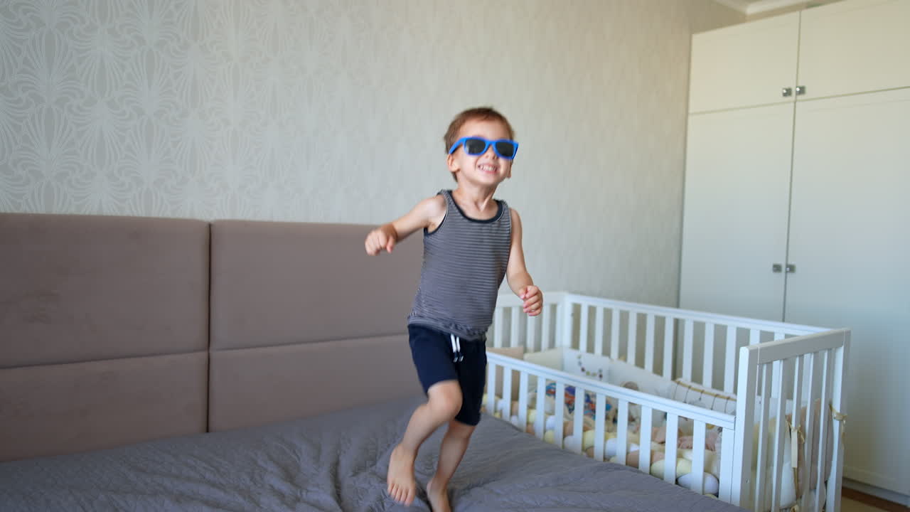 Adorable smiling Caucasian baby puts on his sunglasses. Active kid stands up and jumps on the big bed cheerfully.