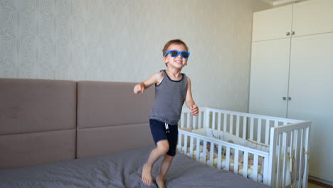 adorable smiling caucasian baby puts on his sunglasses. active kid stands up and jumps on the big bed cheerfully.