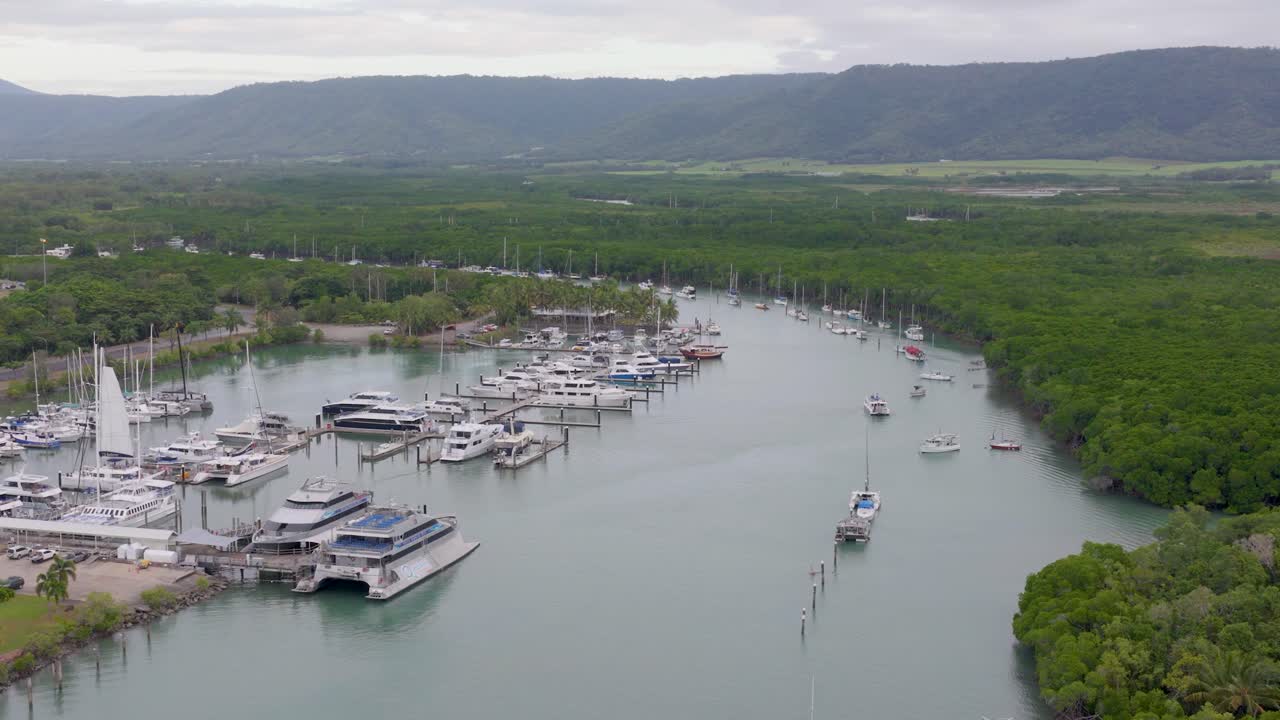 Aerial footage captures boats docked at Port Douglas marina, surrounded by lush greenery and calm waters under overcast skies